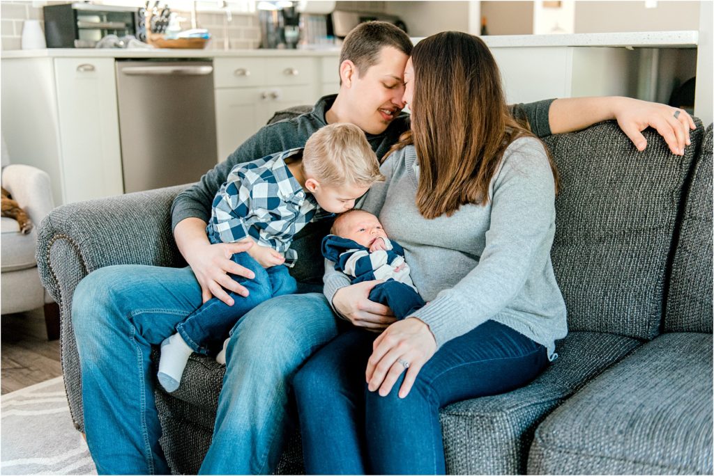 Family snuggling on the couch while big brother kisses his newborn brother's head during an in-home lifestyle newborn photo session.