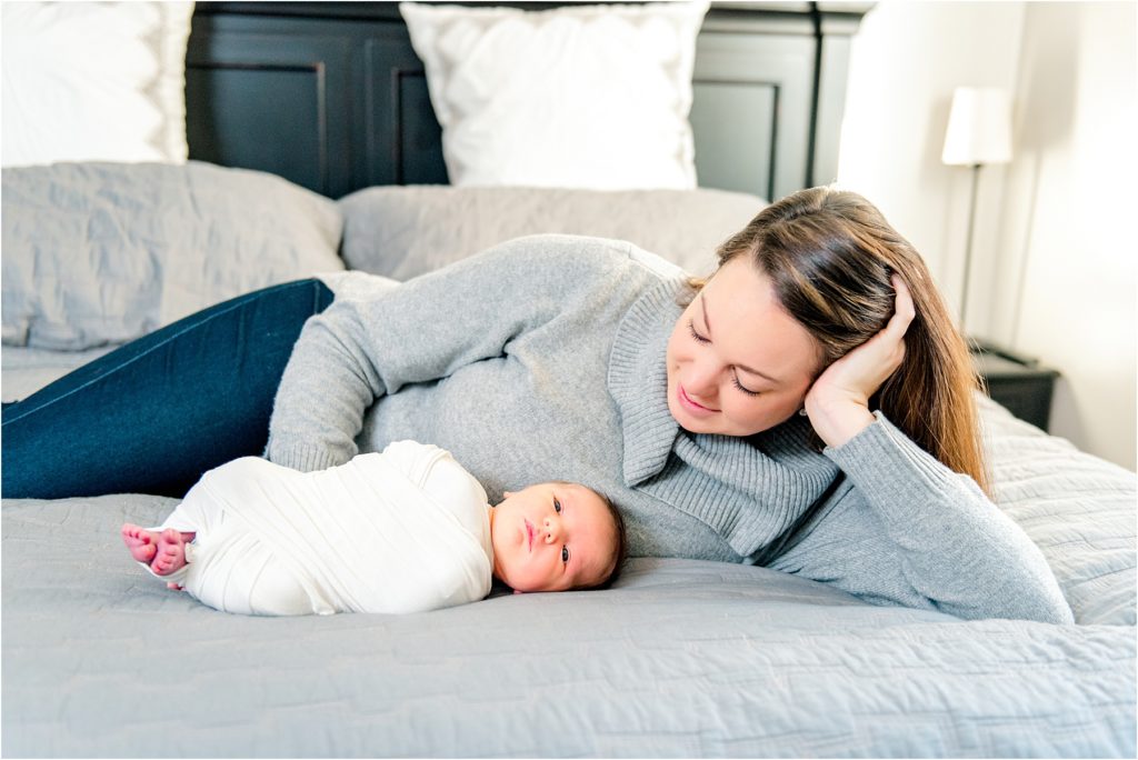Mommy laying behind her newborn baby on her bed, while smiling down at him. In-Home lifestyle newborn photo.