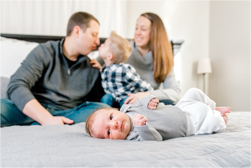 Family photo during an in-home newborn lifestyle photo session. Baby laying on parent's bed while the family is blurred out sitting behind him.