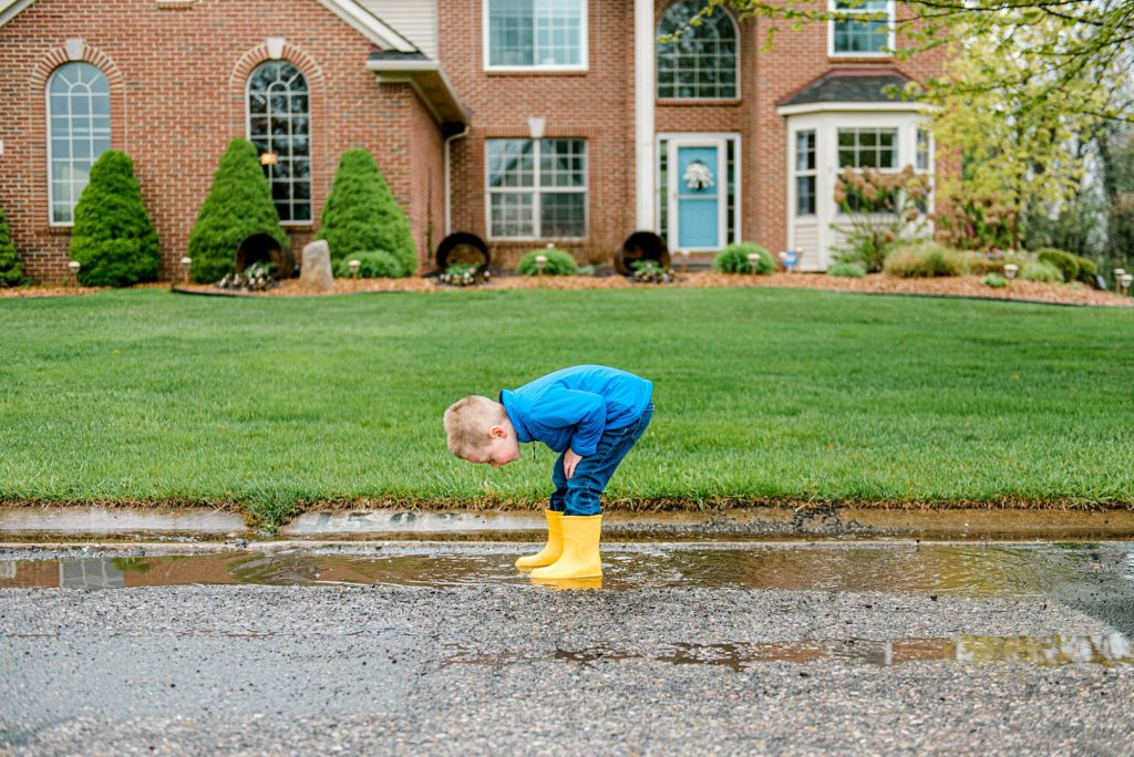 little boy standing in front of his house stomping in rain puddles with yellow rainboots. howell mi photography life lately.