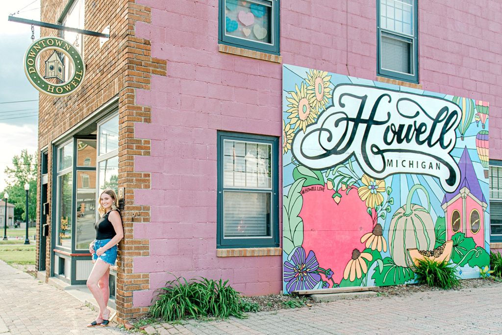 girl standing in front of a pink building with a Howell Michigan mural in Downtown howell.