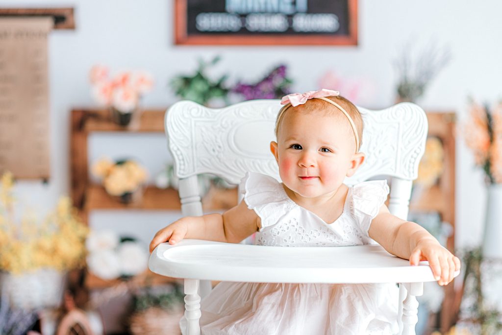 Baby in a pretty vintage white high chair in front of a floral backdrop for her first birthday photos and cake smash.