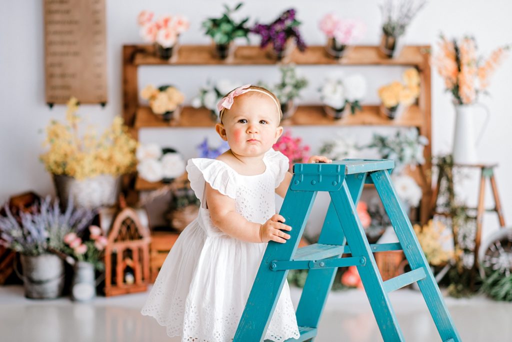Baby holding onto a blue ladder in front of a beautiful floral backdrop 