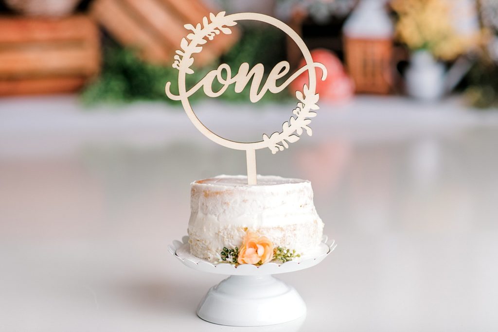 White cake with "One" topper and flowers
