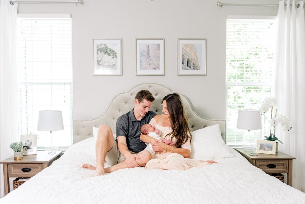 Photo of a mother, father, and newborn baby sitting on the bed in their home, taken by an Ann Arbor Newborn Photographer. 