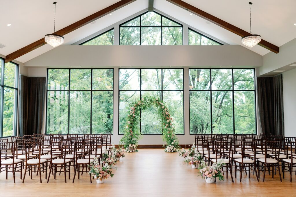 Wedding ceremony site. Brown chiavari chairs and flower arrangements flanking the aisle, with a floral arch at the head of the aisle.