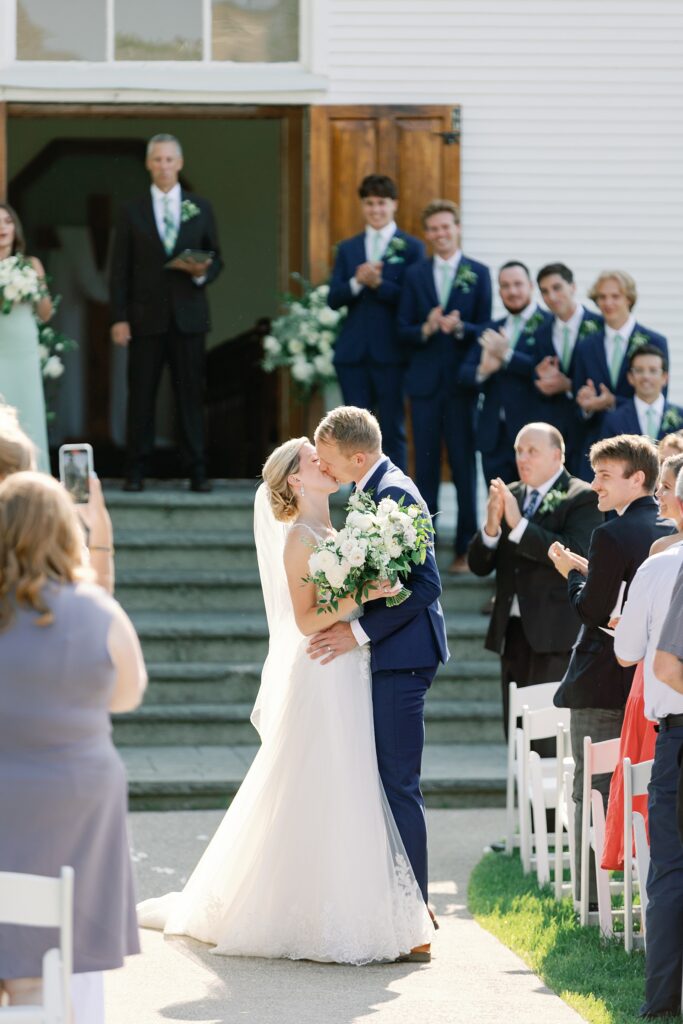 Bride and groom kissing as they walk down the aisle after their ceremony in front of a white chapel at the Felt Mansion